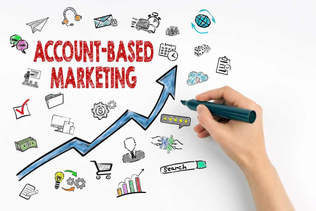 An illustration with the title 'Account-Based Marketing' written in bold red letters in the center. Surrounding the title are various hand-drawn icons representing business concepts, including communication, money, analytics, teamwork, and a rising arrow graph symbolizing growth. A hand holding a marker is seen drawing the upward-trending arrow, with icons like a shopping cart, search bar, and dollar sign integrated into the design, symbolizing different aspects of marketing and strategy.