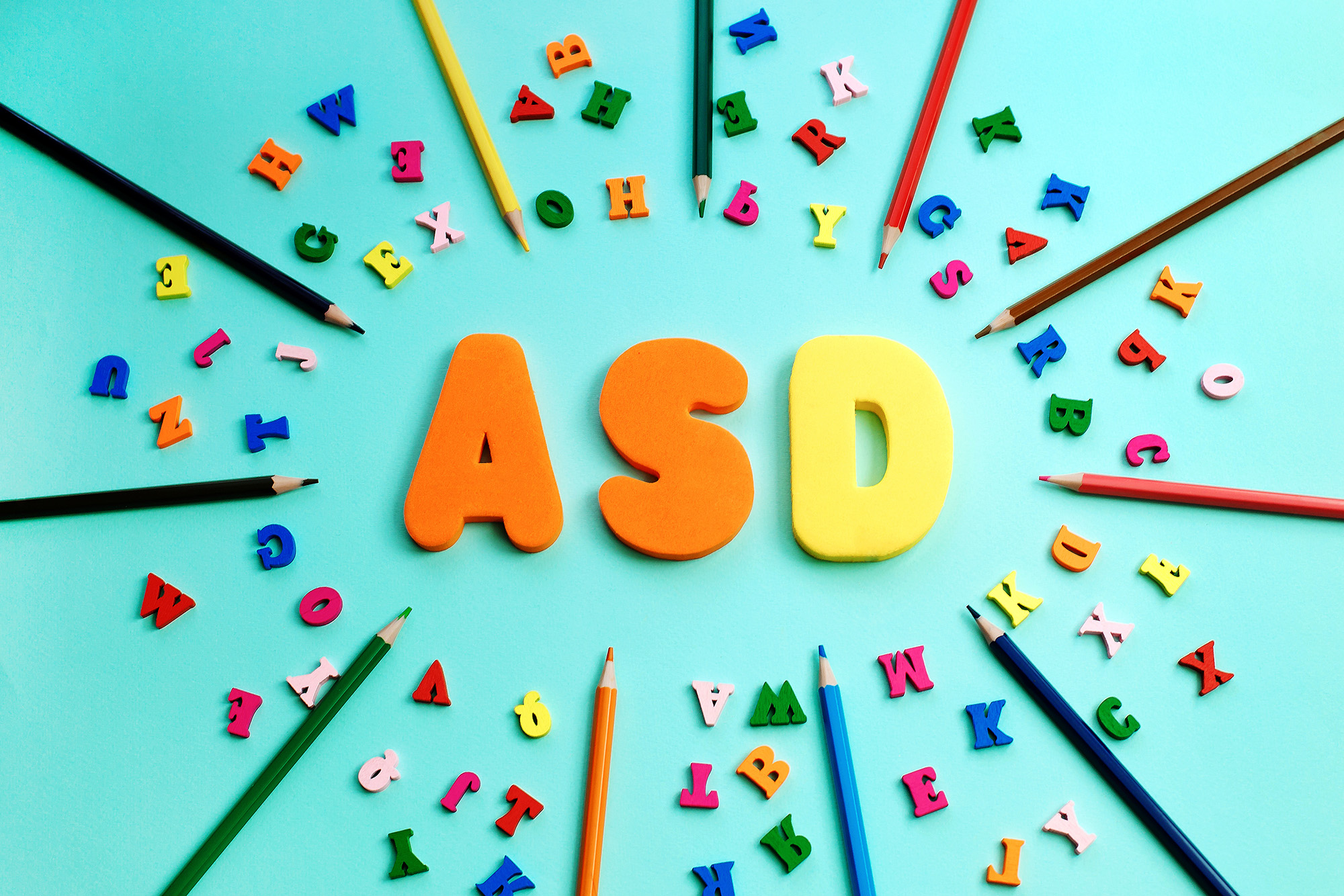 The letters 'ASD' in large, colorful foam block letters are arranged in the center of the image, surrounded by scattered colorful alphabet letters and pencils on a bright teal background. The letters and pencils form a visually vibrant and playful composition, representing creativity and learning.