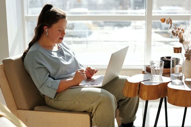 A woman sitting comfortably in an armchair by a window, working on a laptop. She is wearing a light blue top and has earbuds in, appearing focused on her work. A small table beside her holds a glass of water, a few items, and some decorative dried plants. The room is bright with natural light coming through large windows.