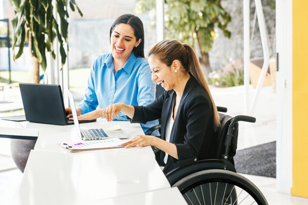 Two professional women working together at a table with laptops, both smiling and engaged in conversation. One woman is wearing a light blue shirt, and the other is in a wheelchair, wearing a black blazer. Both appear to be in a bright, modern office environment with large windows in the background.